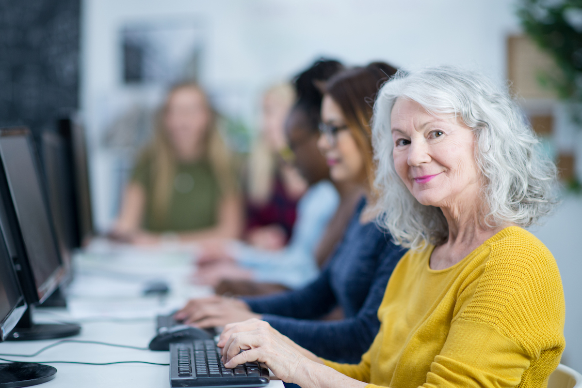 A diverse group of adults are indoors in their university computer lab. A senior Caucasian woman is smiling at the camera while typing on a keyboard.